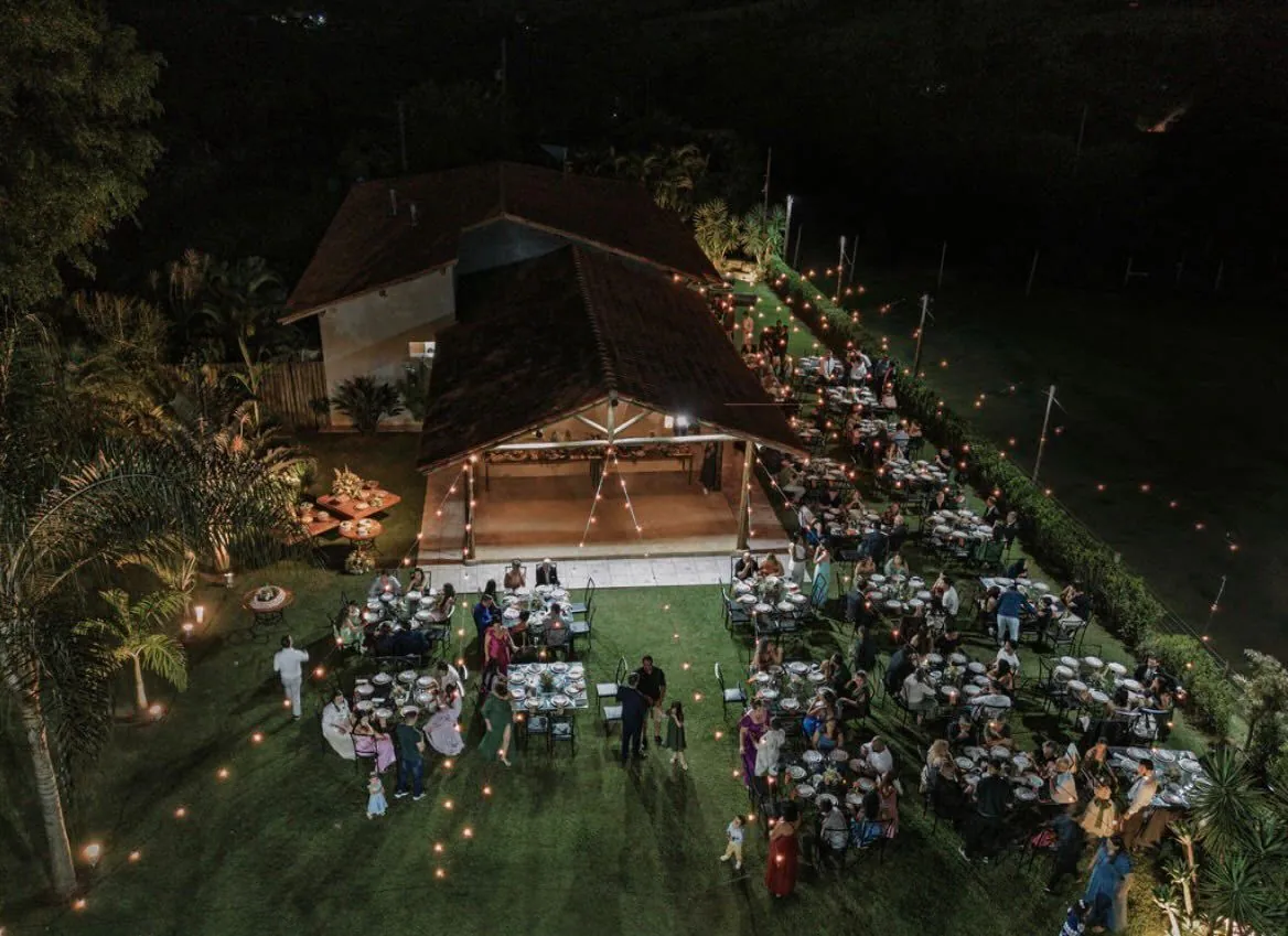 Vista aérea da recepção à noite: salão iluminado, mesas no gramado e caminhos marcados por velas