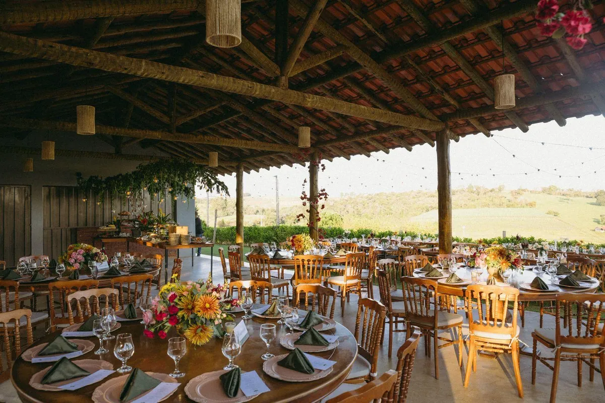 Salão coberto com estrutura de madeira, mesas redondas decoradas e vista para o campo