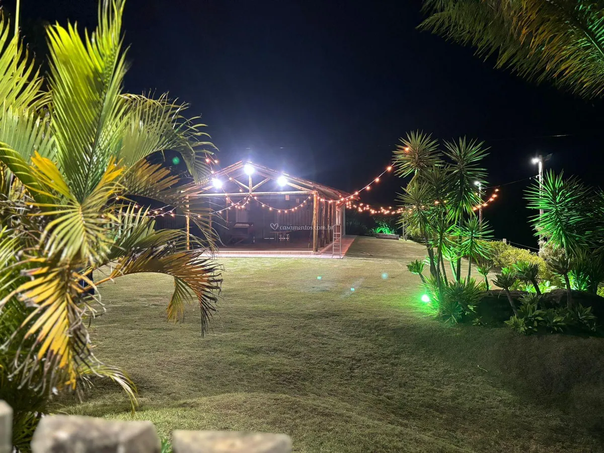 Jardim iluminado à noite, com palmeiras e o salão enfeitado com cordões de luz