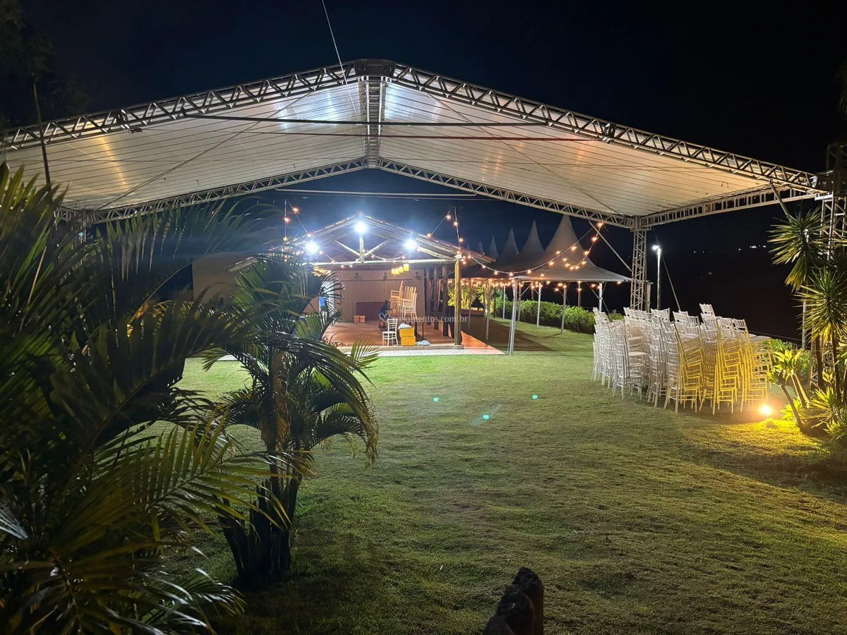 Tenda iluminada à noite sobre o gramado, com o salão e varais de luz ao fundo